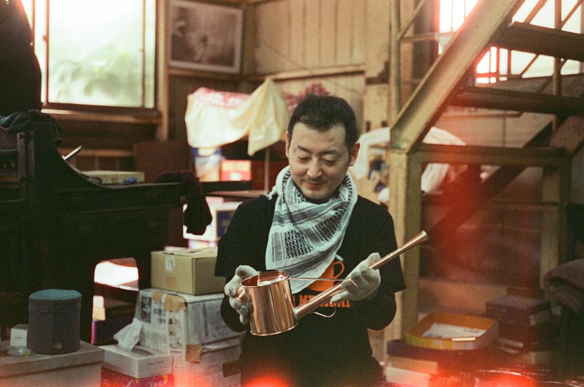 Yoichi Negishi in his workshop holding a copper watering can, the frame washed with a red film light leak